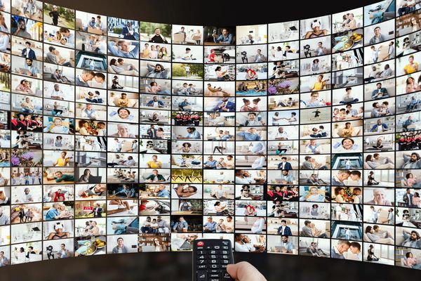 Hand holding remote control in front of a wall of TV screens showing diverse scenes.