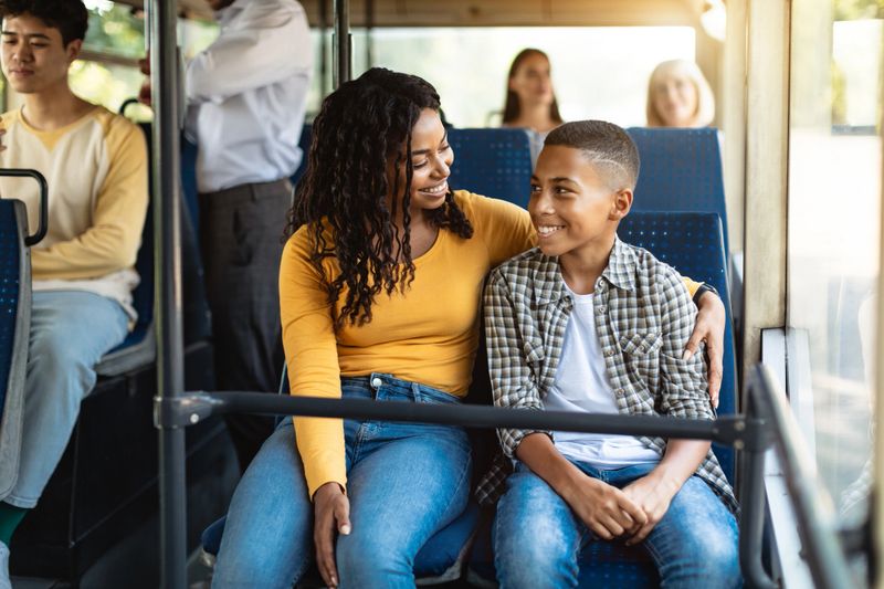 Happy Family On The Bus. Portrait of smiling young African American woman and boy sitting and going on a public transport, lady hugging son and looking at each other, enjoying ride or travel