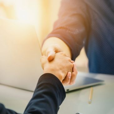 Two people shaking hands over a desk with a laptop and notebook.