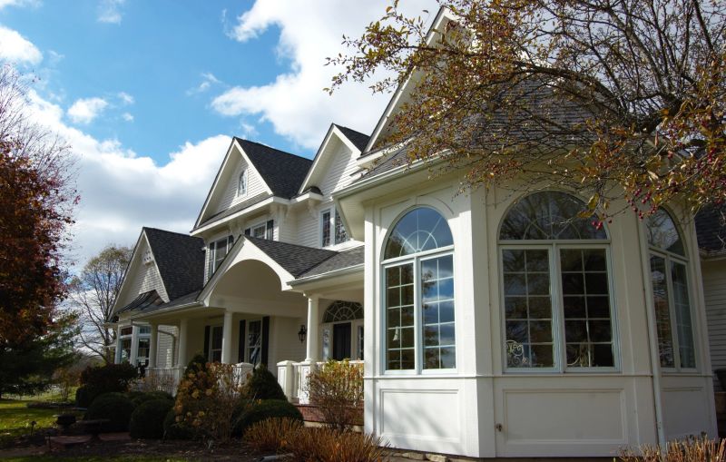 A side angle view of a large, white, victorian style house with landscaping in autumn against a blue sky with clouds.