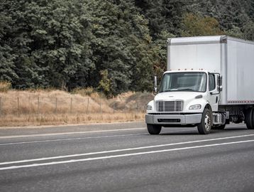 White semi-truck driving on a highway with forest in the background.