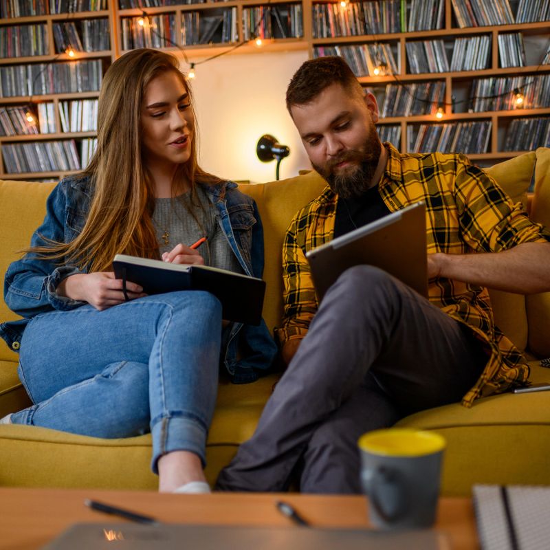 Couple of students using digital tablet and books while studying together at home while sitting on the couch