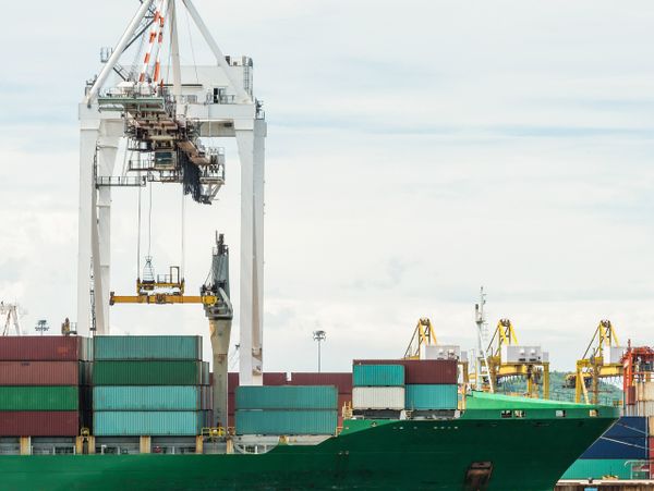 Green cargo ship docked at port with containers and cranes.