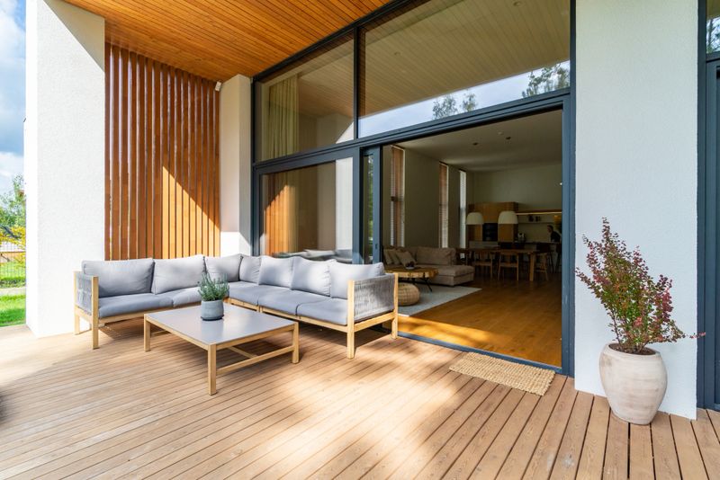 Wooden patio connected to a private building, with seating area and cozy lounge zone, open window with entrance to the house.