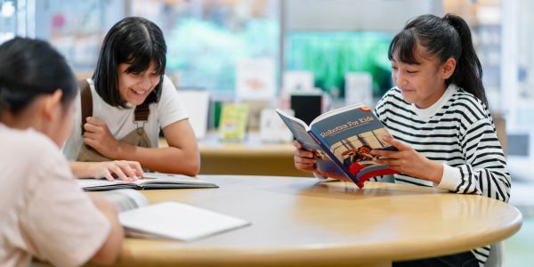Three kids reading books at a round table in a library.