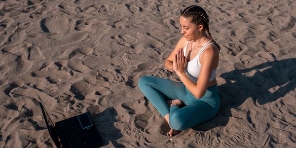 Woman practicing yoga on sandy beach with laptop nearby.