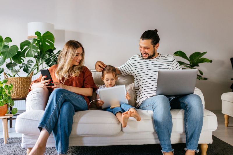 Married diverse couple and little preschool daughter sitting on couch using computer watching movie cartoons online surfing internet shopping purchasing via internet spending weekend together at home