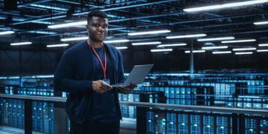 Man holding laptop in a high-tech data center with blue-lit servers.