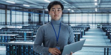 Man in a gray turtleneck holding a laptop in a large data center.