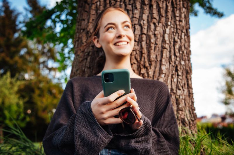 Happy smiling young woman leaning on tree in the garden, reading messages on her mobile phone, looking up, smiling happy towards the camera. Young women social media lifestyle concept shot.