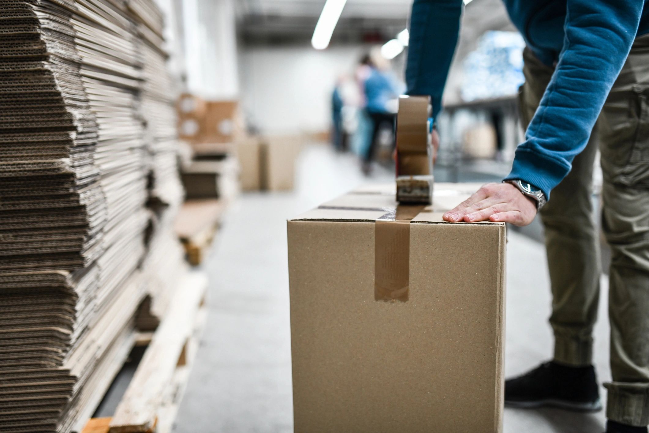 Person sealing a cardboard box with tape in a warehouse.