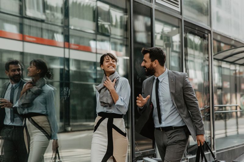Spontaneous photo of a handsome bearded young adult male, a businessman wearing a gray suit, walking outdoors the glassed working building, and talking with his working partner, a brunette with bangs, stylishly dressed. Both having a positive conversation, looking happy