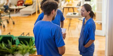Healthcare workers in blue scrubs talking in a bright room with patients relaxing in the background.