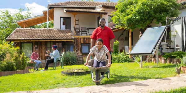 A man pushes a child in a wheelbarrow in a sunny garden with people chatting in the background.