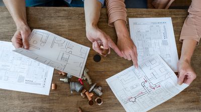 Two people reviewing architectural blueprints and plumbing parts on a wooden table.