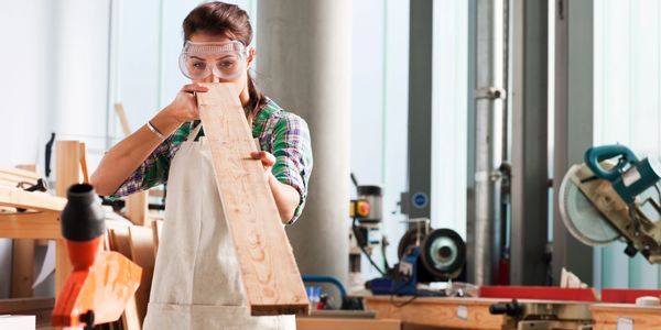 Woman inspecting a wooden plank in a workshop wearing safety goggles and apron.