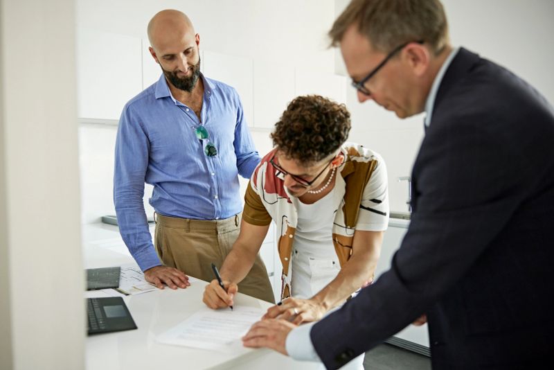 Salesman in suit watching as mid adult gay couple begins process of signing documents to finalize purchase in modern kitchen of their new home.