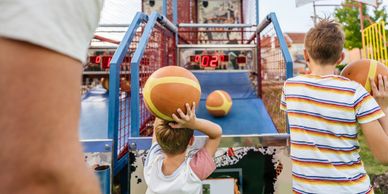 Two boys playing basketball arcade game outdoors with a man watching.