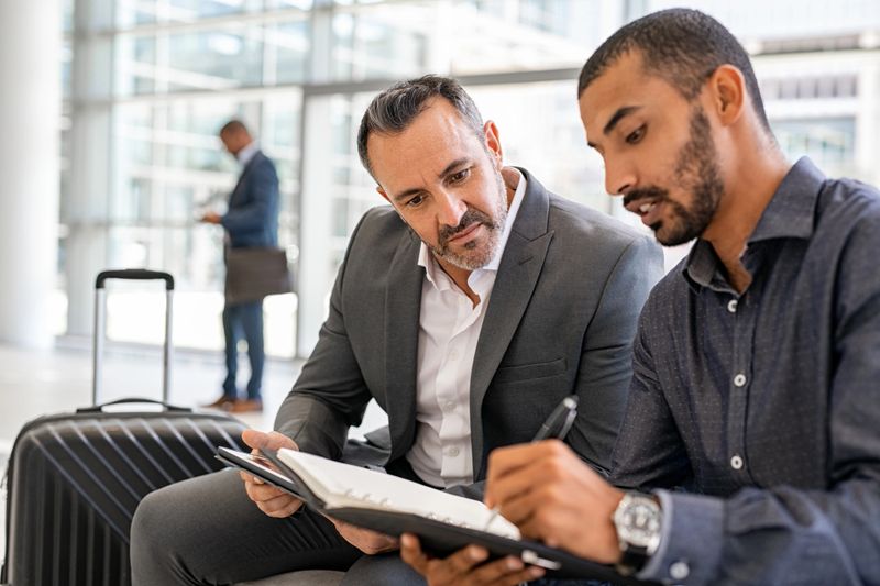 Two multiethnic businesspeople sitting at departure lounge at the airport discussing work while making notes in agenda. Indian business man working at airport with worried manager while waiting in lounge their connection. Successful businessman discussing during a business travel.