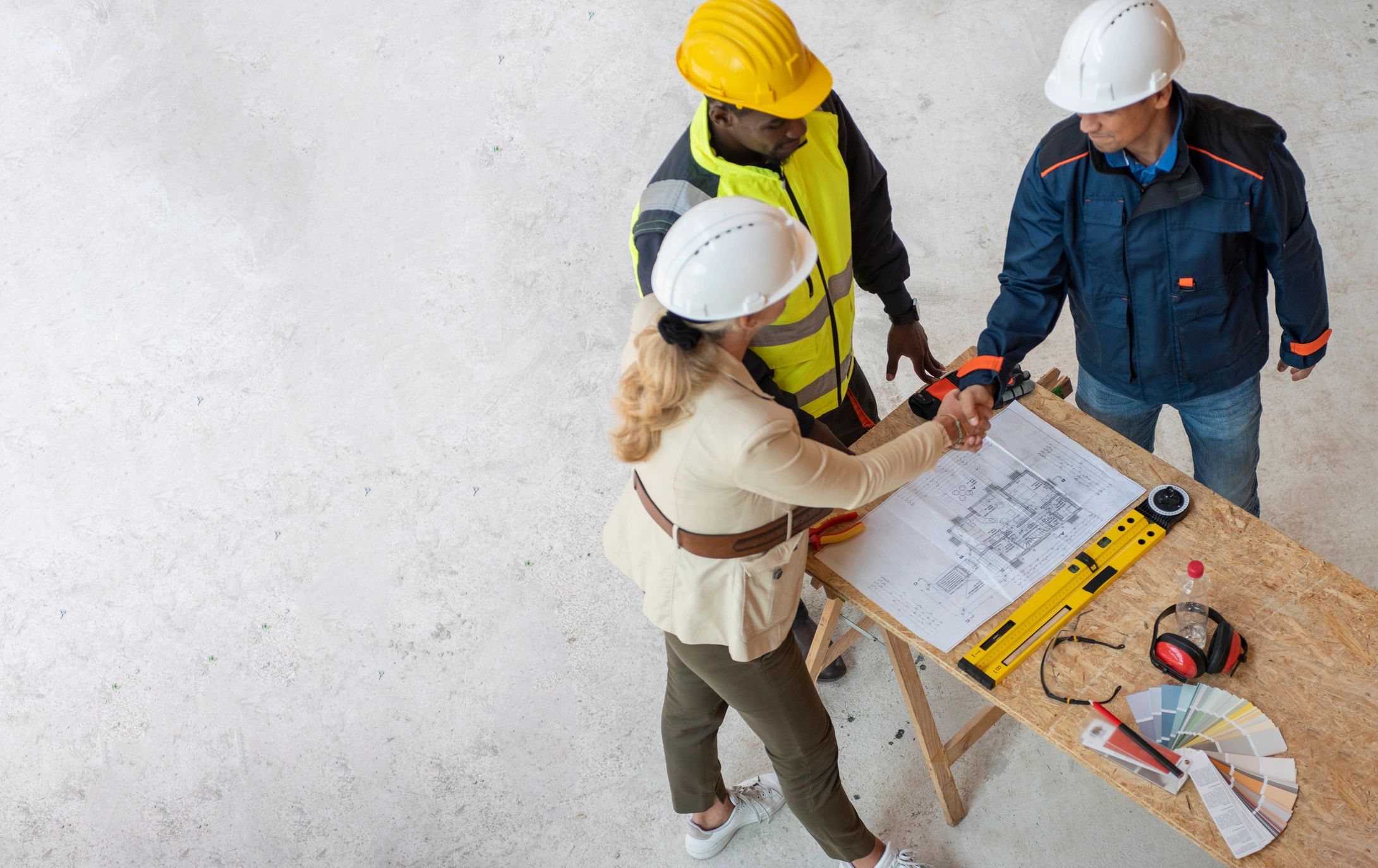 Two construction workers shaking hands over a table with blueprints and tools.