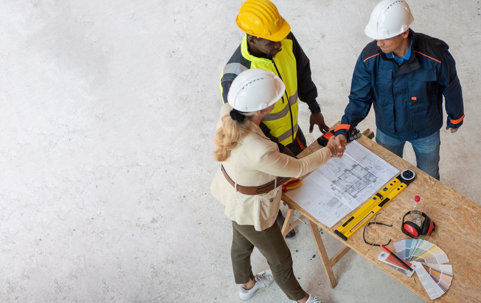Two construction workers shaking hands over a table with blueprints and tools.