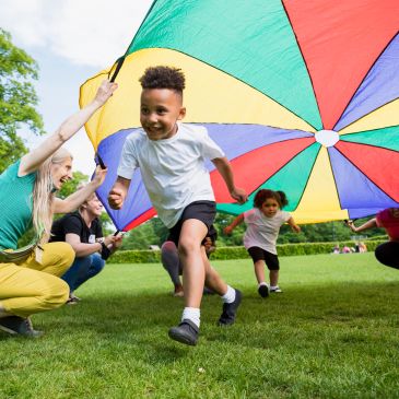 Children running under a colorful parachute held by adults outdoors.