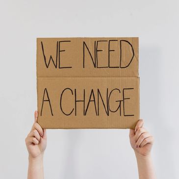 Hands holding a cardboard sign saying 'WE NEED A CHANGE'.