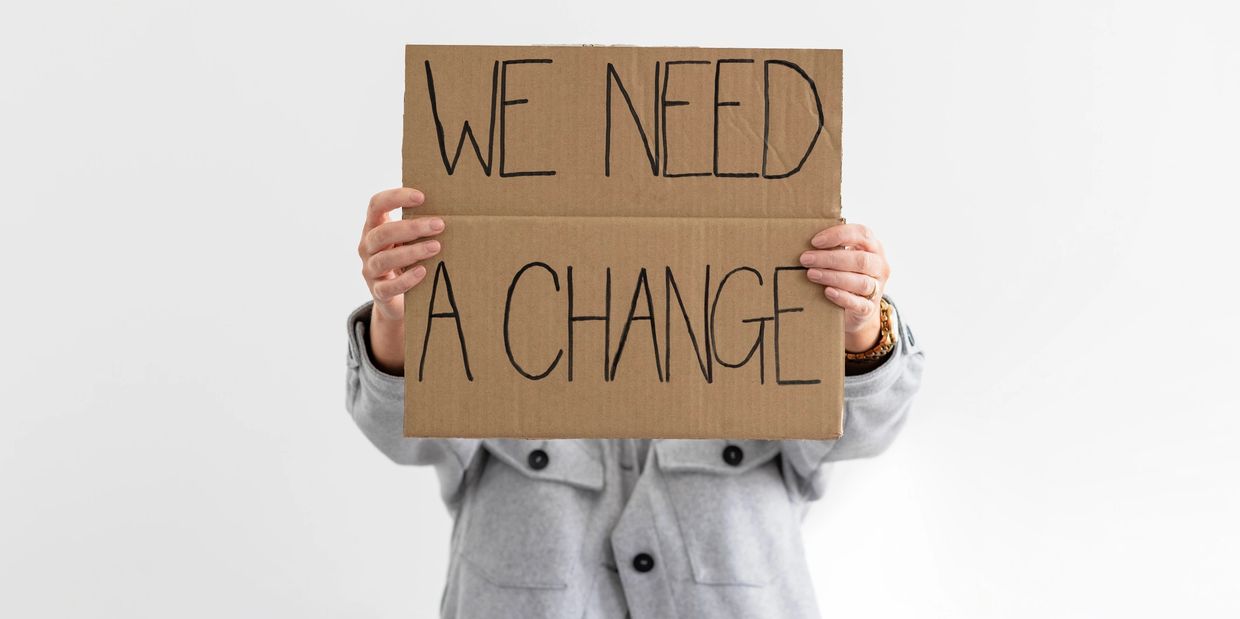 A sign that reads, "We need a change" held in front of the face of a person wearing a grey shirt.