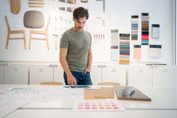 Man working on design plans in a workshop with fabric samples and tools.