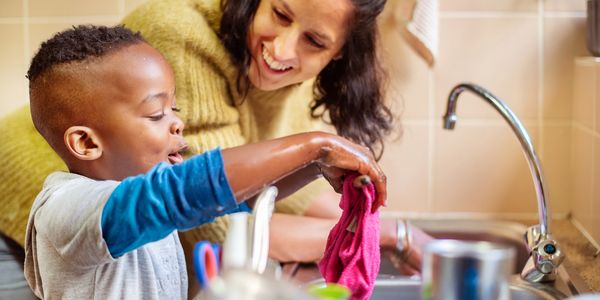 A child and woman happily washing dishes together at a kitchen sink.