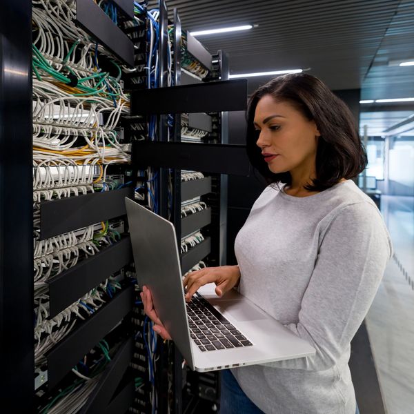 Woman using laptop in a server room with network cables.