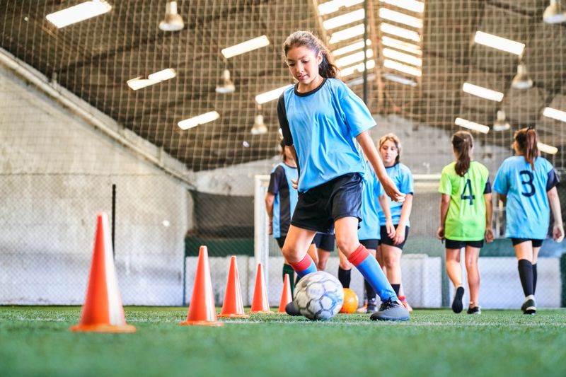 Young girl leading ball between cones on sports court. Female player in blue jersey practicing dribbling skill at court.