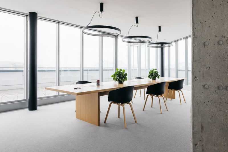 A photo of a spacious office with modern furniture and large windows. In the middle is a large wooden desk with chairs. No people in the photo. Horizontal daylight indoor photo.