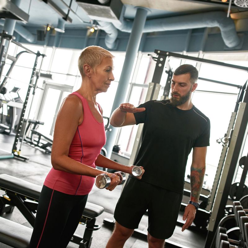 Side view fit mature woman with short blond hair standing with dumbbells in gym next to supportive trainer. Professional instructor carefully helps to perform physical activities. Active lifestyle.