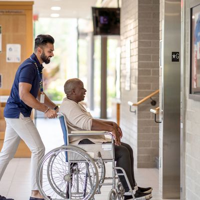 A young man pushes an elderly man in a wheelchair in a hallway.