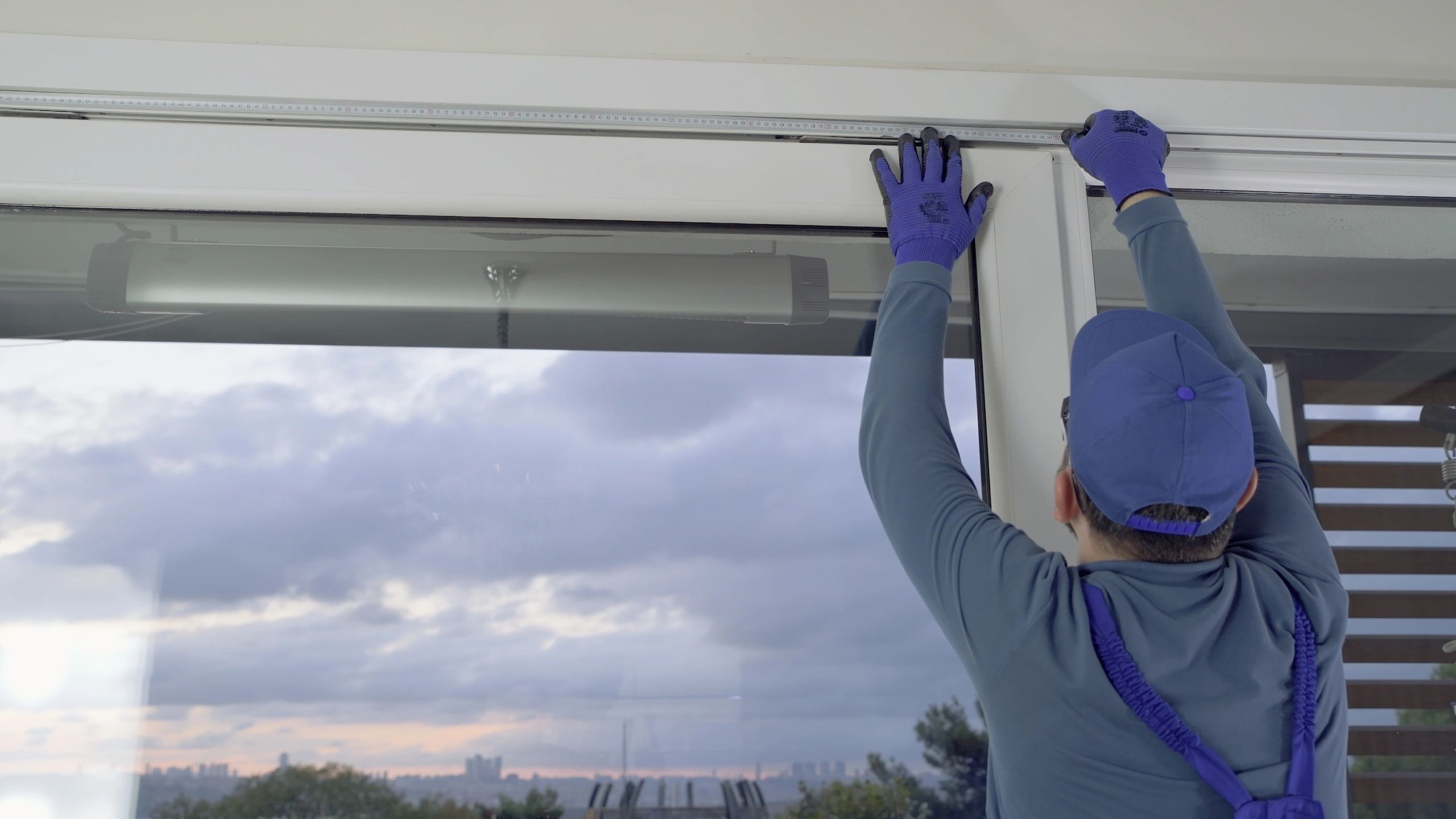 A worker in blue gloves and cap measures a window frame indoors.