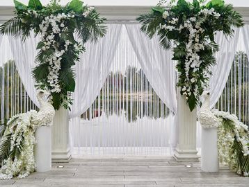 Elegant wedding arch decorated with white flowers and peacock sculptures beside a lake.