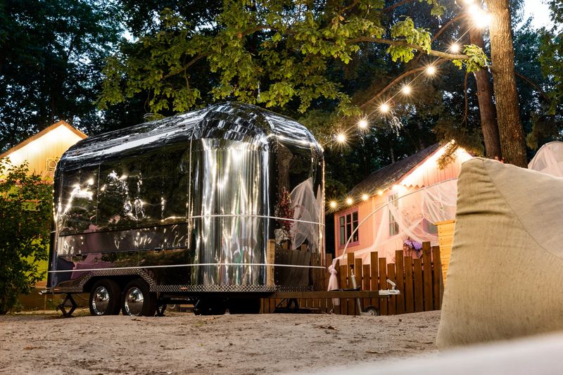 Scenic evening view of vintage futuristic silver metal camper van trailer parked near wooden fence at countryside farm or ranch against light garland forest tree. Retro style fodtruck caravan vehicle.