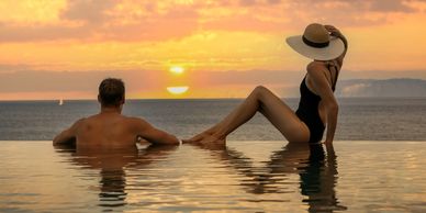A couple relaxes in a pool watching a vibrant sunset over the ocean.