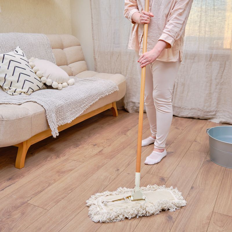 An adult woman mops a wooden laminate floor while cleaning her home living room.