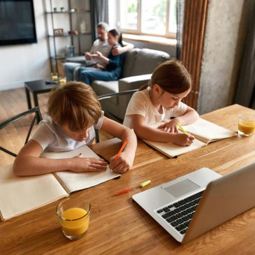 Two children focused on homework at a wooden table with orange juice and a laptop.