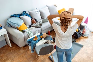 Woman overwhelmed by clutter in her living room, holding her head in frustration.