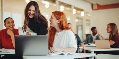 Three young women happily collaborate around a laptop in a modern classroom.