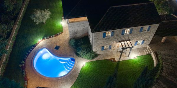 Night aerial view of a stone house with a lit swimming pool and garden.