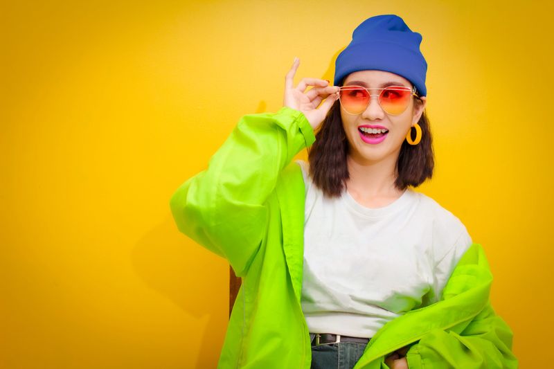 Smiling of happy young woman 20 years holding glasses-she wearing a green jacket and white shirt with blue jeans fashion cloth enjoy sitting on chair on yellow background