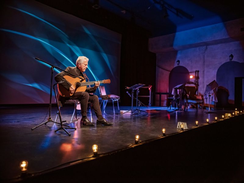 Mature male Guitarist on the stage with classic guitar. Grey hair. He is wearing all black. Stage interior of Theatre during performance at night.
