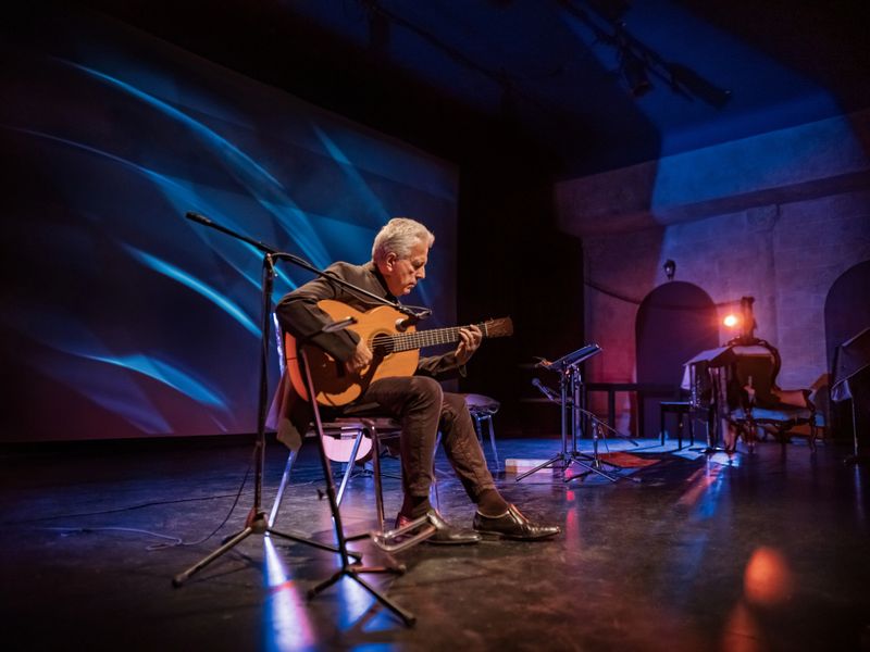 Mature male Guitarist on the stage with classic guitar. Grey hair. He is wearing all black. Stage interior of Theatre during performance at night.
