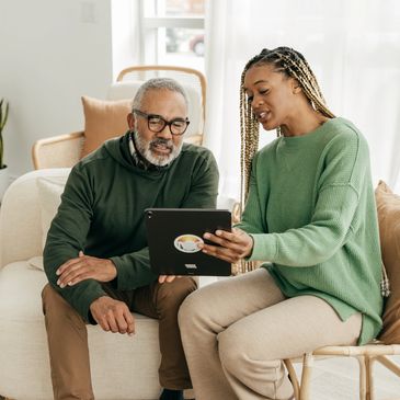 A woman shows a tablet to an older man in a cozy living room.