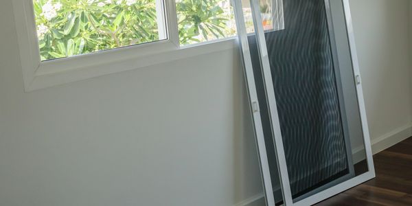 Two window screens leaning against a wall near a window with green foliage outside.