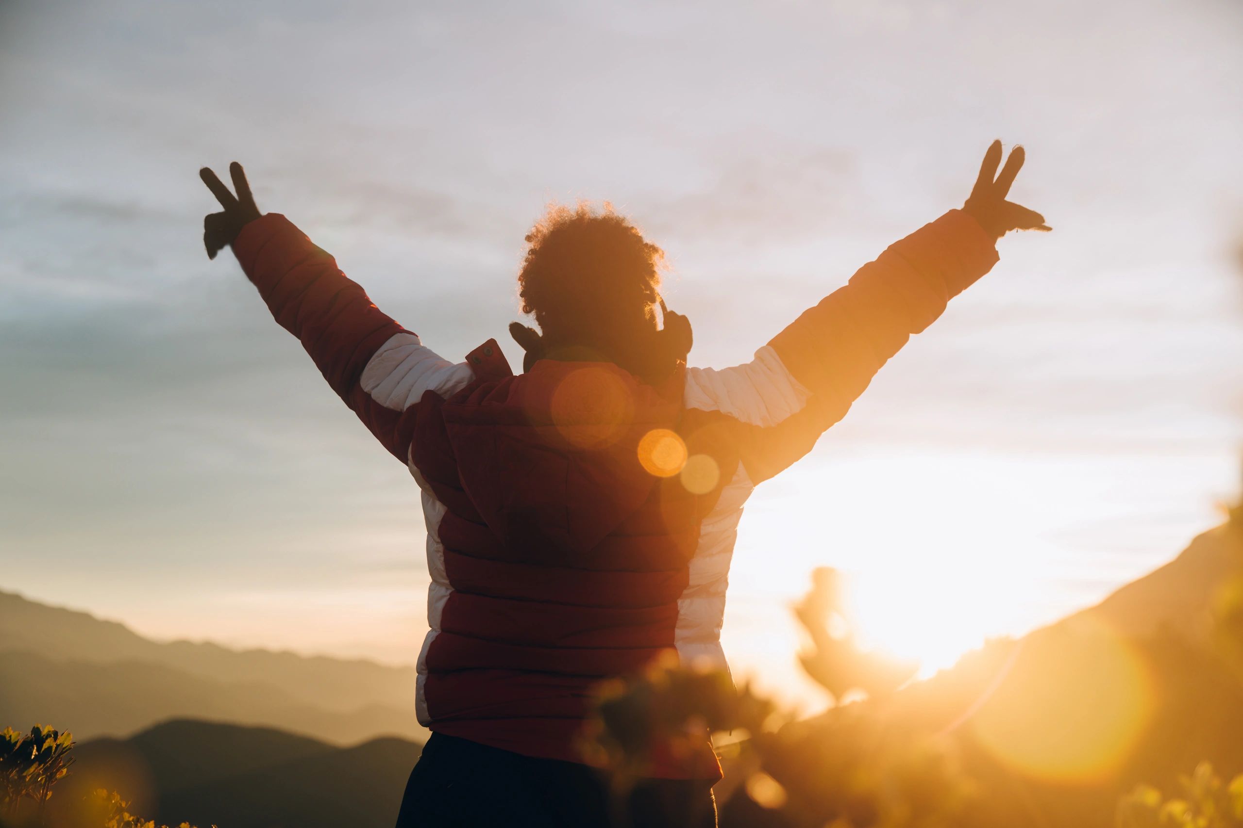 Person in a warm jacket raising hands in peace signs at sunset outdoors.